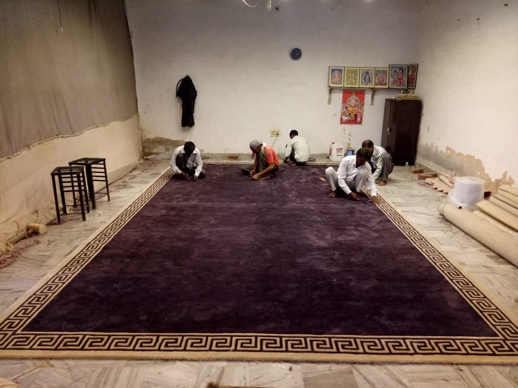 Handmade purple carpet manufacturing in Udaipur, skilled artisans stitching custom rug border for hotels and halls.